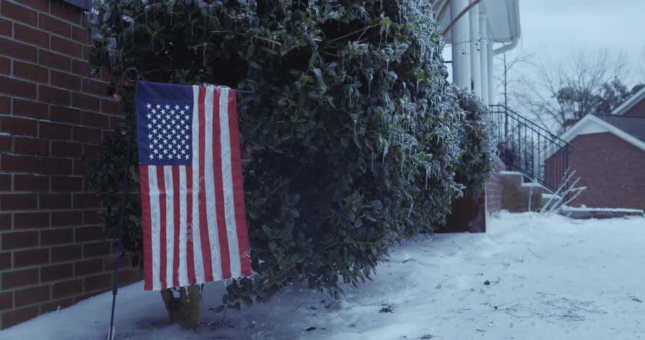 An American flag stands proudly in a snow-covered front yard next to ice-laden bushes after a winter storm