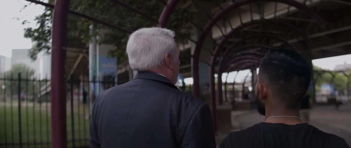 Two men walk past a fence by a bridge