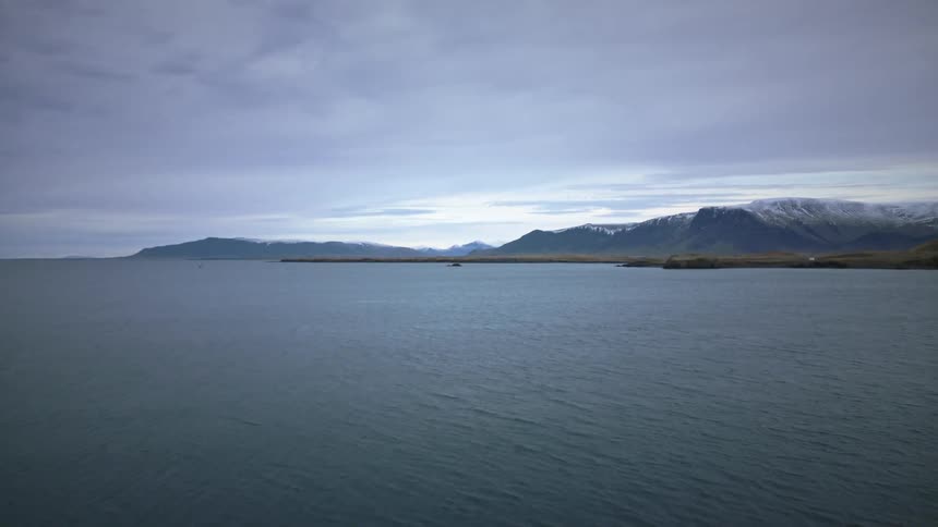 A serene view of an Icelandic fjord with gentle waters flowing towards distant, snow-capped mountains under a cloudy sky
