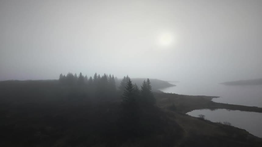 Slow aerial footage pushes in on a moody, fog-shrouded landscape with a small evergreen forest on the shore of a tranquil lake
