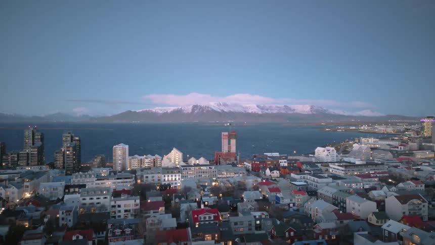 A slow, pulling-back aerial shot reveals the glowing cityscape of Reykjavik, Iceland, with the ocean and snow-capped mountains at twilight
