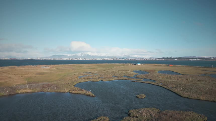 Serene aerial footage reveals a vast coastal wetland, with a distant city and snow-capped mountains across the water under a clear blue sky