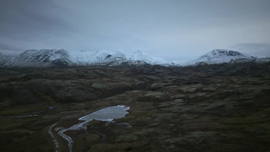 A slow pull-back shot reveals a vast, rugged landscape with a frozen lake and snow-capped mountains in the background under an overcast sky