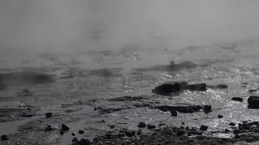 Steam rises from a bubbling geothermal hot spring in a stark, primordial landscape