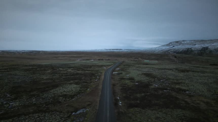 A high-angle aerial view slowly rises over a desolate road in a vast, moody landscape with snow-covered mountains on the horizon