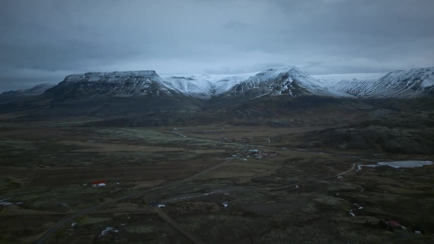 A dramatic wide aerial shot reveals a vast Icelandic landscape with a small village resting in a valley below epic snow-capped mountains