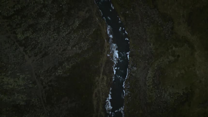 A moody top-down aerial view slowly ascends over an icy river flowing through a dark, mossy Icelandic landscape