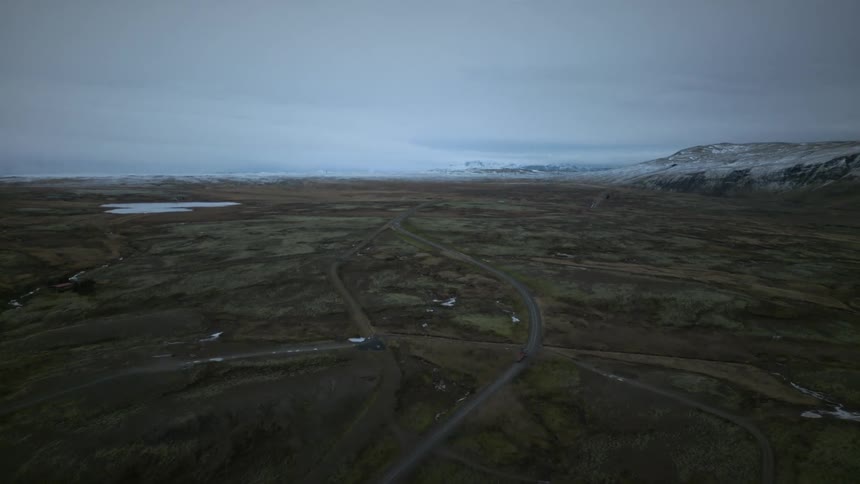 An epic aerial view captures vehicles traveling along a winding road through a vast, remote, and mossy Icelandic landscape towards distant snow-capped mountains