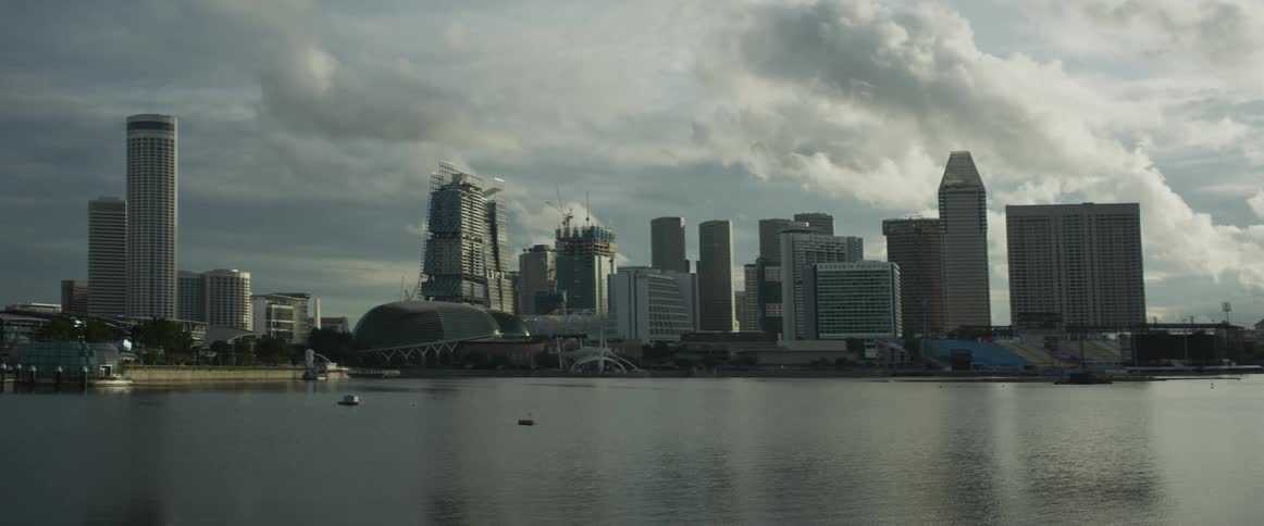 A wide shot shows the Singapore skyline and the Esplanade across the calm bay on a cloudy day