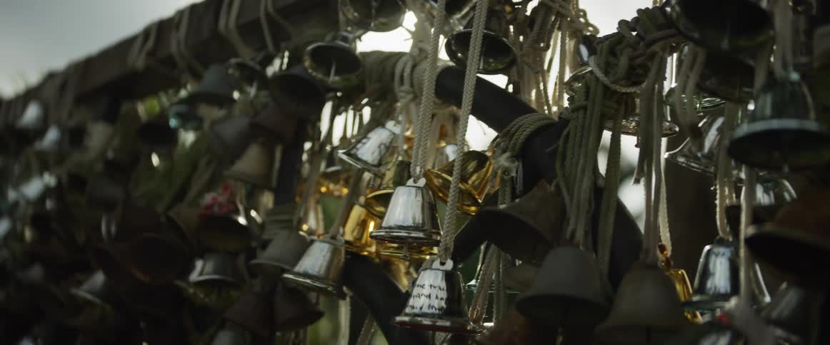 A close-up slow-motion pan reveals a dense collection of silver, gold, and bronze bells hanging from ropes in the sunlight