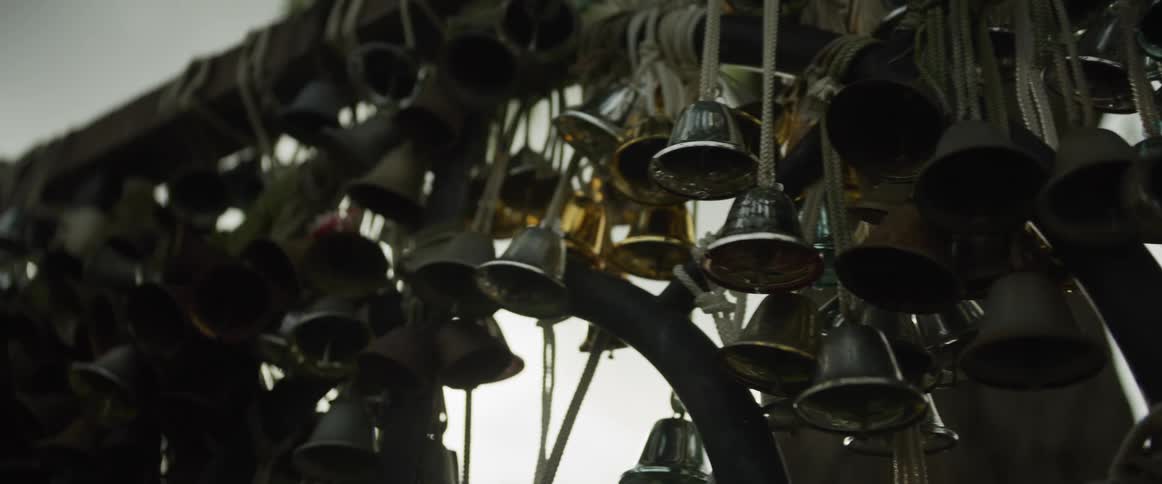 A collection of small, assorted metal bells hang from ropes as the camera slowly pans across with sunlight peeking through