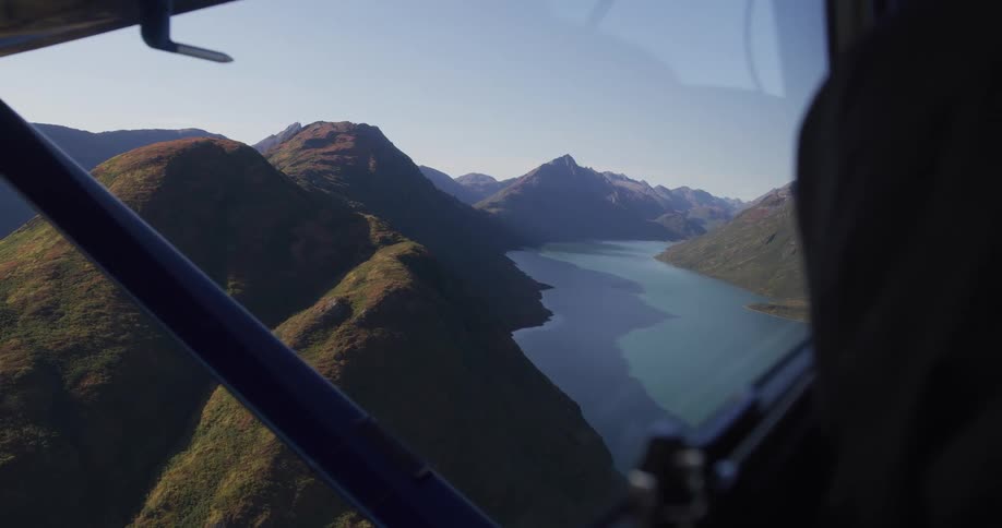 An aerial point-of-view from a small plane flying over a majestic mountain range and a winding lake in the Alaskan wilderness on a clear day