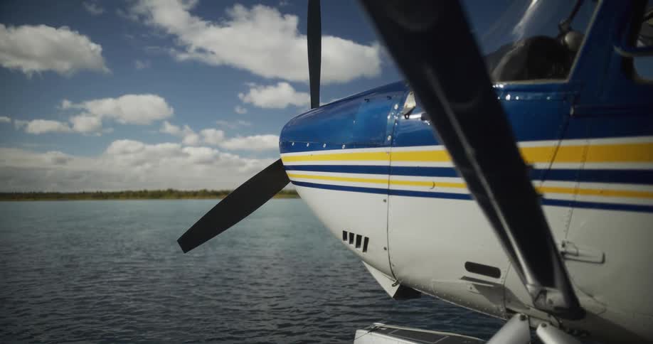 A still seaplane floats peacefully on a lake, with a close-up on the propeller under a partly cloudy sky