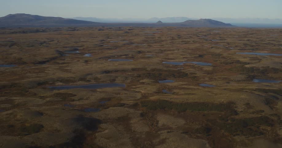 A wide, static aerial view shows the vast, remote tundra landscape dotted with numerous small lakes, with mountains visible on the distant horizon