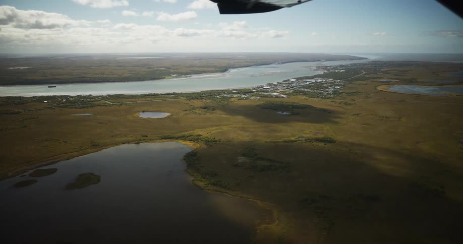 A stunning aerial view from a small plane shows a remote town nestled along a wide river in a vast, watery tundra landscape