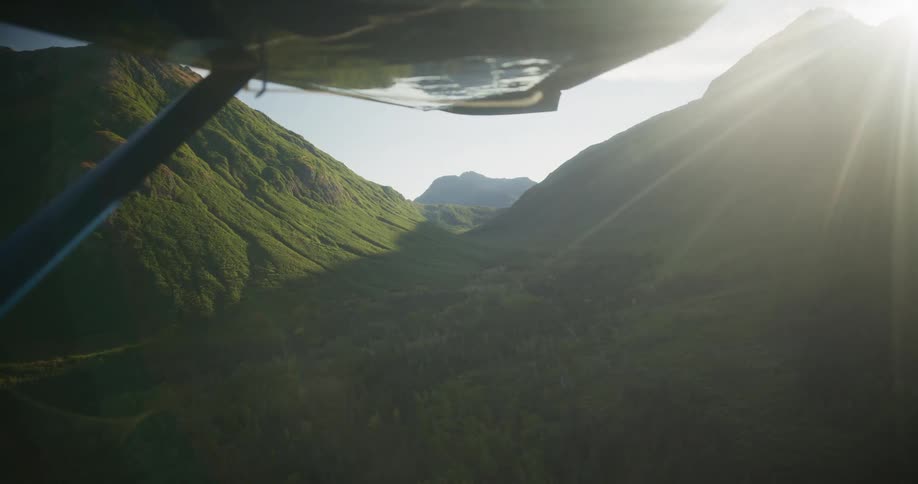 An aerial view from under the wing of a small plane flying over a lush green mountain valley as the sun rises