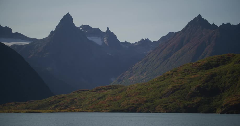 A tranquil alpine lake sits before rolling green hills and a majestic, rugged mountain range with snow-capped peaks