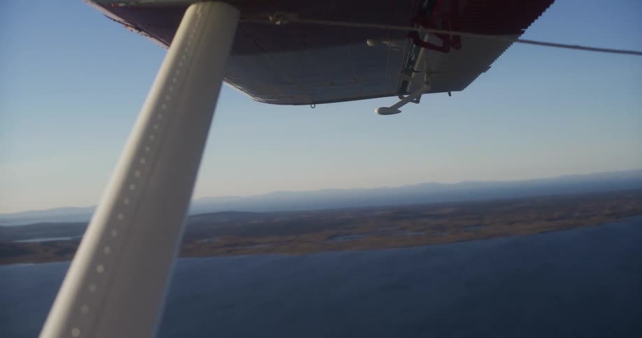 View from a small plane window showing the wing while flying over a scenic coastline and ocean on a clear day