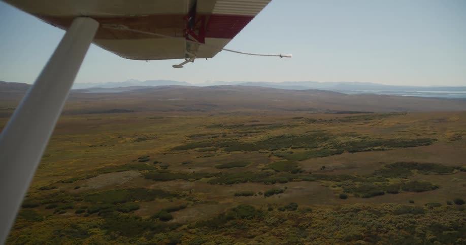 An aerial view from under the wing of a small plane reveals the vast, remote tundra of Alaska stretching towards distant mountains and the ocean