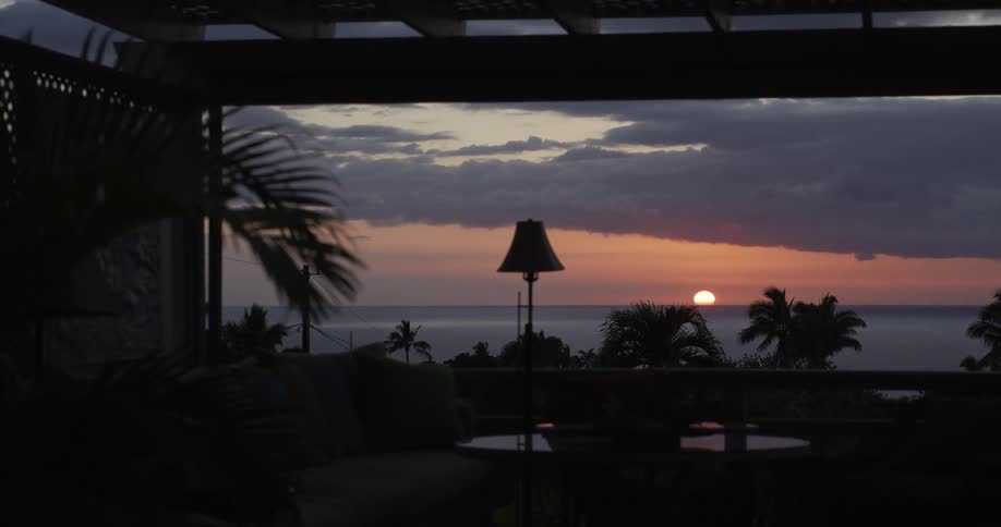 A tranquil sunset dips below the ocean horizon, viewed from a silhouetted tropical patio with palm trees