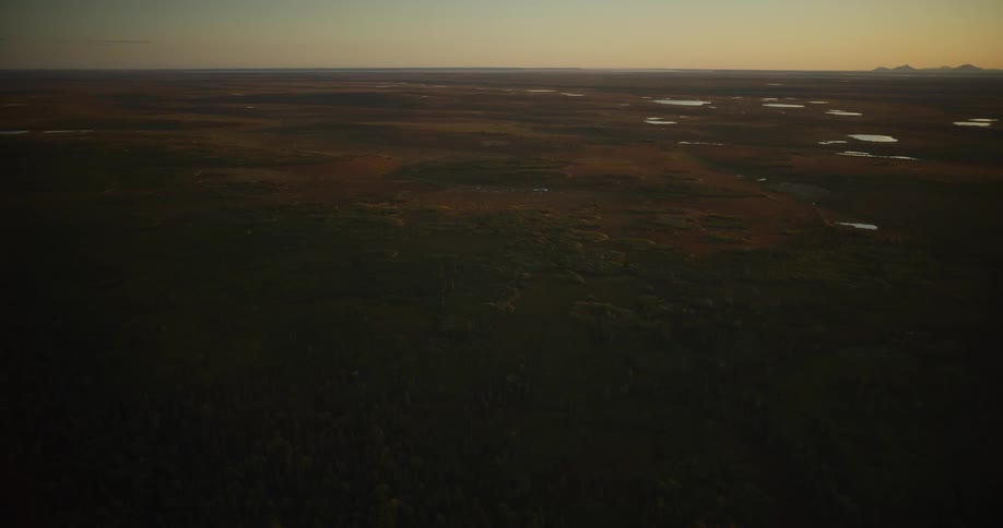 A slow aerial shot flies over a vast northern landscape of tundra, forest, and scattered lakes during a golden sunset