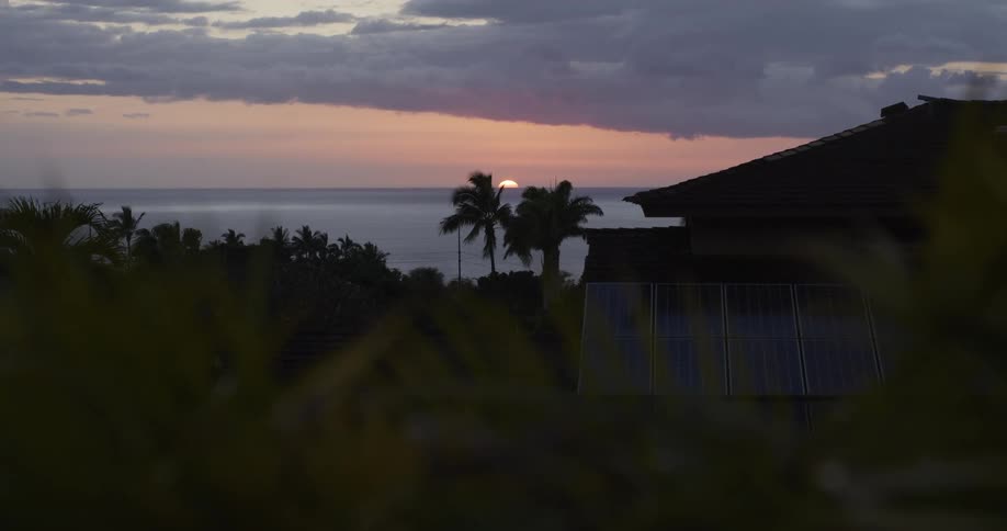 Peaceful sunset over the ocean with a home featuring solar panels and palm trees in the mid-ground