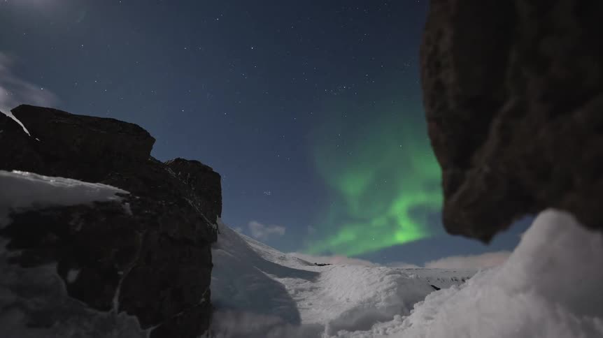 A stunning timelapse of the vibrant green Aurora Borealis dancing across the moonlit, starry sky above a snowy mountain landscape