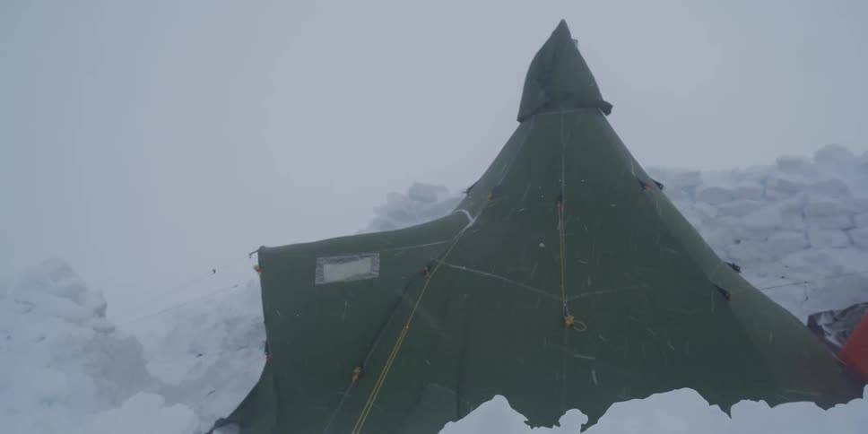 A green expedition tent stands firm against the whipping wind and heavy snow of a blizzard