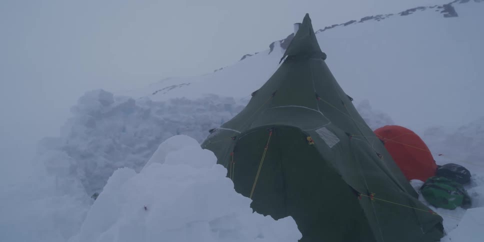 A large teepee-style tent endures a powerful blizzard while pitched on a snow-covered mountain