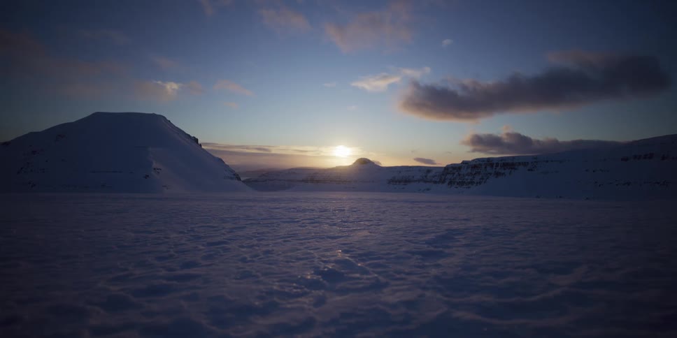 The low winter sun casts a warm glow over a vast, frozen valley between snow-covered mountains