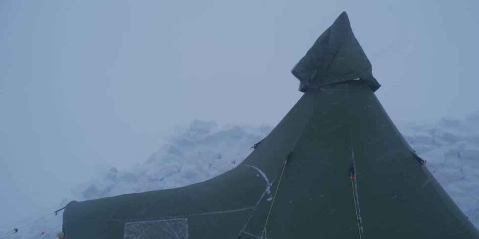 A large green teepee tent stands firm against a powerful blizzard in a harsh, icy environment