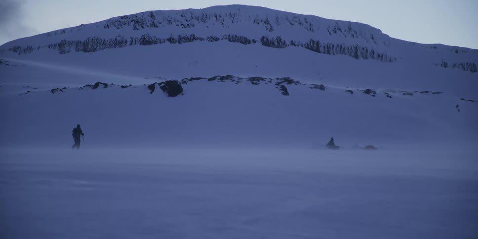 A silhouetted hiker battles a snowstorm while trekking across a vast, frozen expanse at twilight