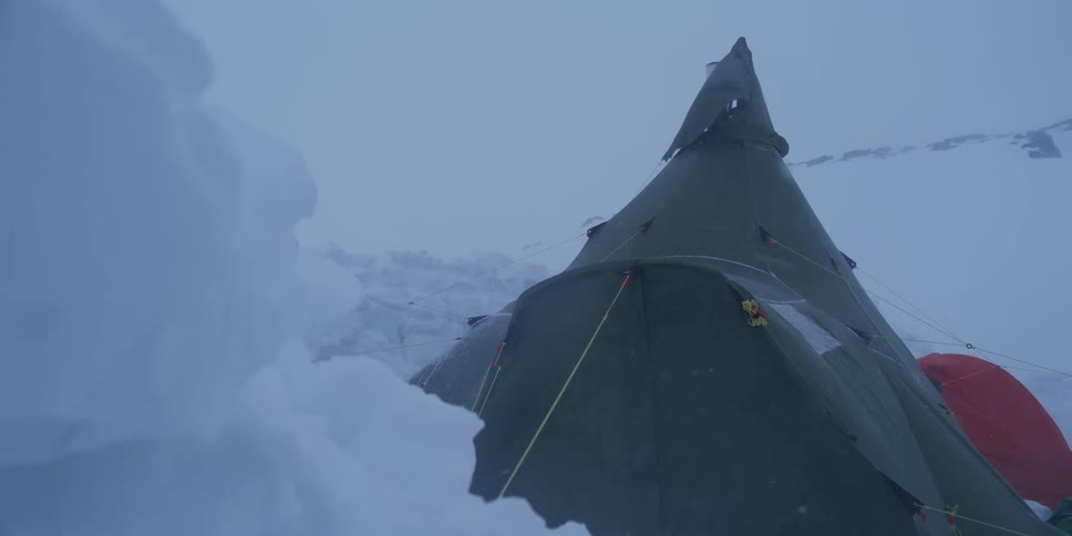 A green, tipi-style hot tent endures a fierce blizzard in a vast, snow-covered arctic wilderness