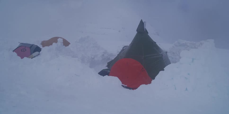A mountain base camp with colorful tents weathers a fierce blizzard, partially buried in deep snow