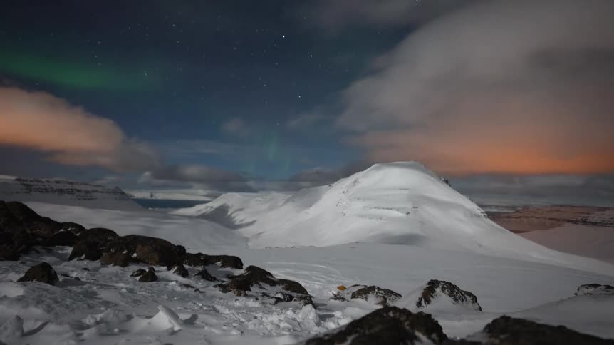 A stunning timelapse captures the ethereal dance of the Aurora Borealis and moving clouds over a vast, moonlit snowy mountain landscape
