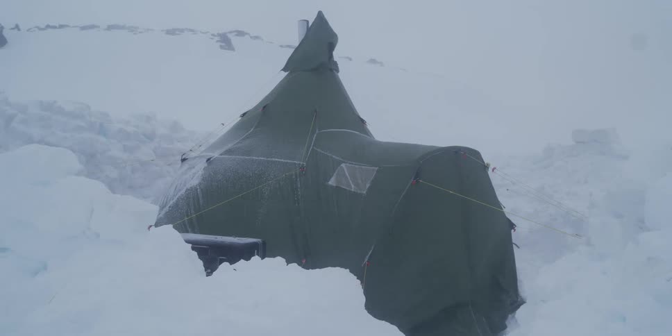 A large expedition tent is buffeted by strong winds during a fierce blizzard in a remote, snowy landscape