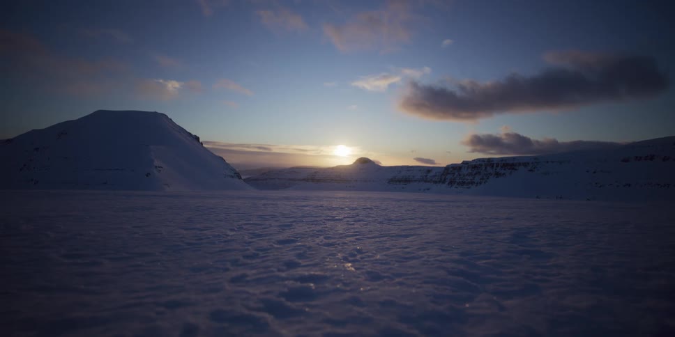 The sun sits low on the horizon over a vast, serene snow-covered mountain landscape