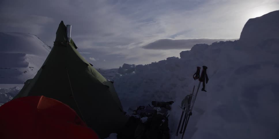 A camera slowly pulls back to reveal a remote winter campsite with a heated tent and ski gear in a vast, snowy landscape