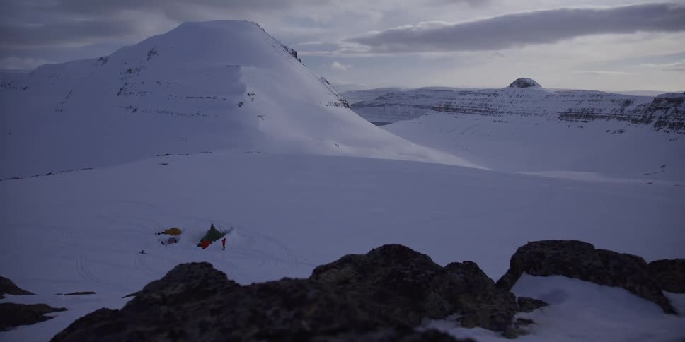 A remote campsite with tents is nestled in a vast, snow-covered mountain valley under a cloudy sky