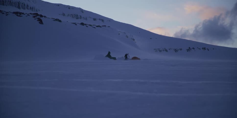 An arctic adventurer sets up camp with a shovel during a snowstorm in a vast, frozen landscape at dusk