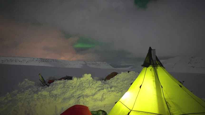 A timelapse shows the Northern Lights dancing in the night sky above an arctic campsite with a glowing tent as clouds and a snowstorm pass over