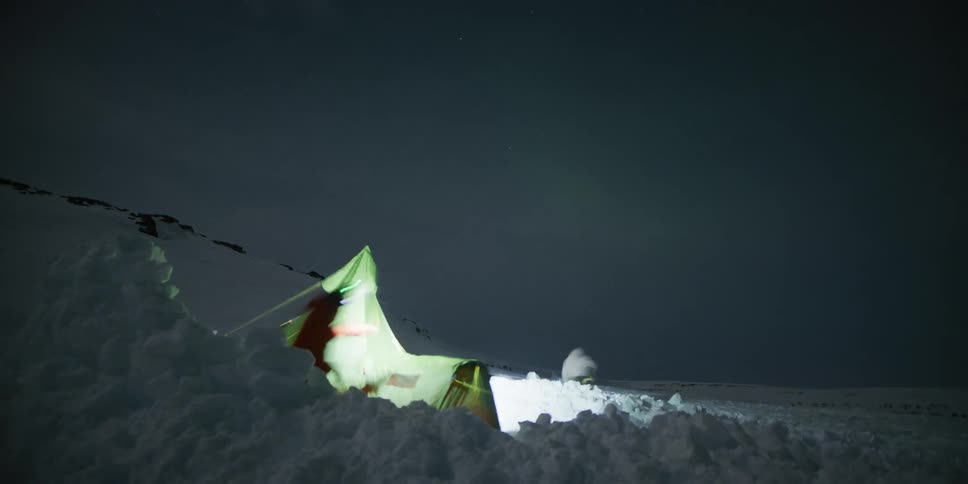 Time-lapse of mountaineers camping in a harsh, snowy landscape at night as the aurora borealis glows faintly in the sky