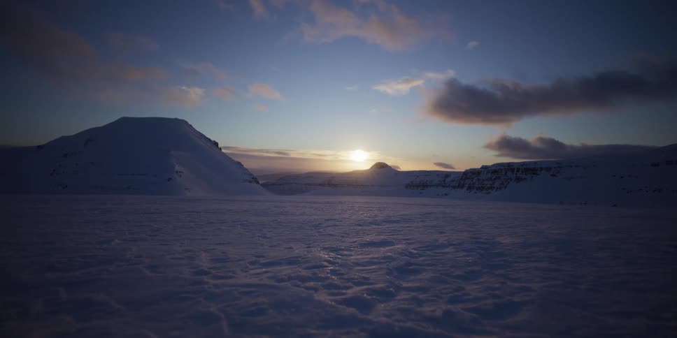 A serene sunrise casts a warm glow over a vast, windswept snowy landscape surrounded by mountains