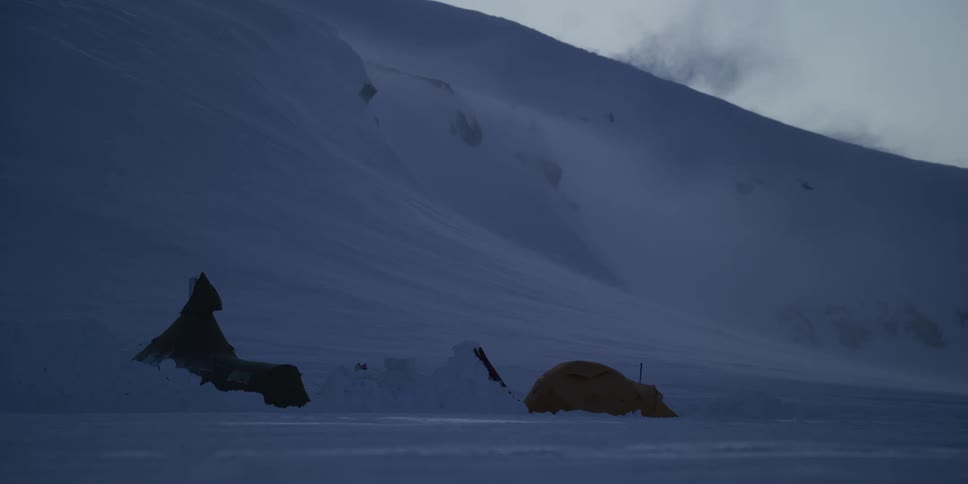 Tents pitched on a snowy mountainside are battered by strong winds during a blizzard at dusk