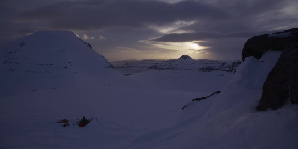 A remote campsite sits in a vast, snow-covered valley surrounded by mountains under a dramatic, cloudy sunset