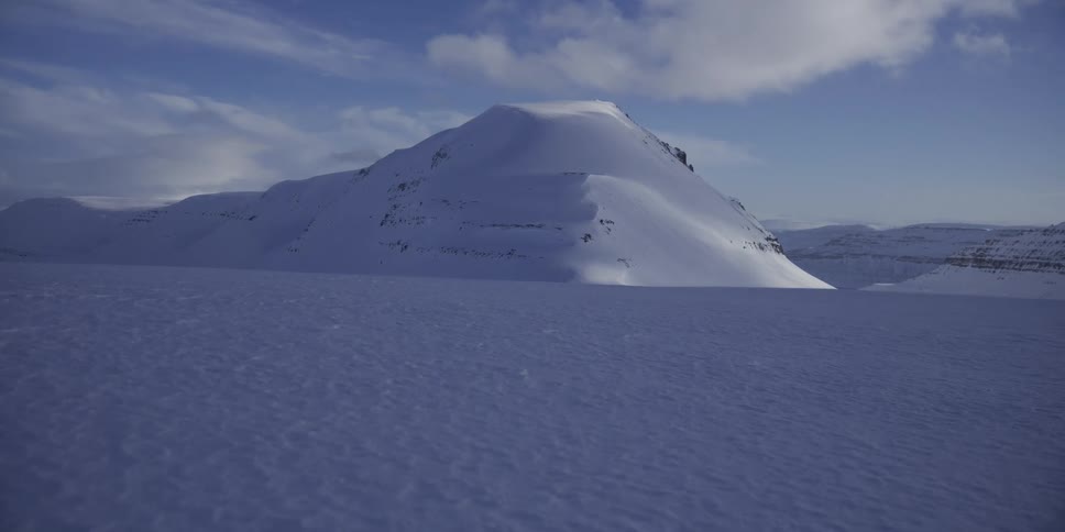 A still, wide shot captures a tranquil and vast snow-covered mountain landscape in the arctic on a sunny day