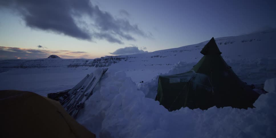 A quiet arctic expedition campsite with tents pitched in deep snow amongst the vast frozen mountains at twilight