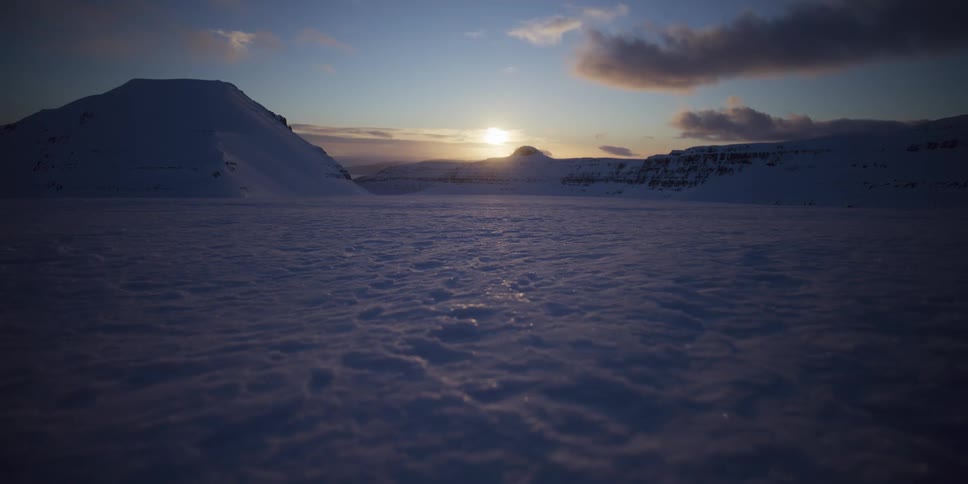 A slow pull-back shot over a vast, pristine snow-covered valley with mountains silhouetted by the setting sun on the horizon