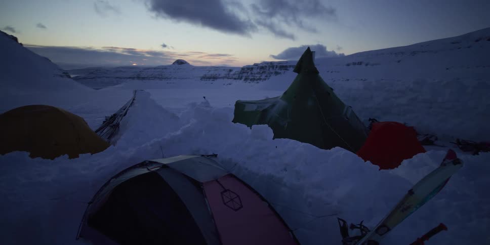 A remote arctic campsite with tents pitched in deep snow amidst a vast mountain landscape during twilight