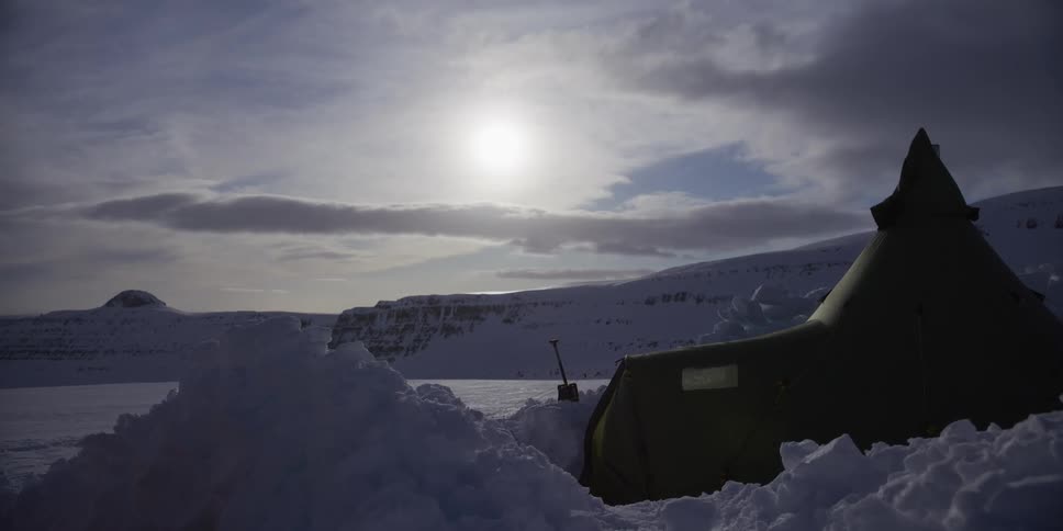 A remote arctic campsite with tents pitched in deep snow under the low-hanging sun and a vast, cloudy sky